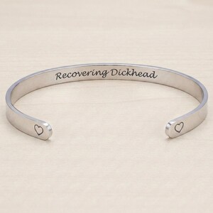 Peut inclure: Un bracelet jonc en métal argenté avec une finition lisse et polie. Le texte "Recovering Dickhead" est gravé en police cursive noire au centre, et un petit contour de cœur simple est gravé à chacune des deux extrémités ouvertes.
