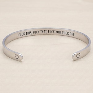 May include: A silver-coloured metal cuff bracelet with an open design. The interior surface is engraved with the black text FUCK THIS, FUCK THAT, FUCK YOU, FUCK OFF. Each end of the bracelet features a small, simple black outline of a heart symbol.