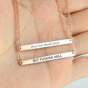 May include: A rose gold-toned double bar necklace featuring two horizontal metal bars suspended from delicate chains. The top bar is engraved with the text this too shall pass, and the bottom bar is engraved with the text BUT FUCKING HELL.
