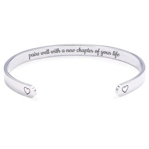 May include: A silver-colored cuff bracelet with the engraved text "pairs well with a new chapter of your life." The bracelet has a heart shape engraved on each open end. Jewelry.