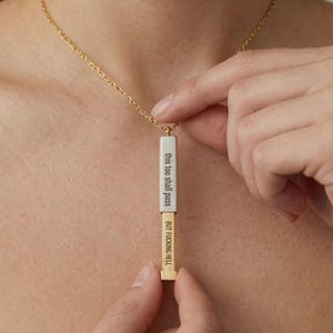 May include: Gold-tone necklace with a rectangular pendant. The pendant is divided into two sections, one white and one gold, with the phrases "this too shall pass" and "BUT FUCKING HELL" printed in black. The necklace is being held by a person.