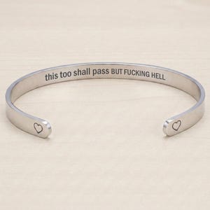 May include: A silver cuff bracelet with the engraved text "this too shall pass BUT FUCKING HELL." Small heart shapes are at each open end. The bracelet is made of a shiny, metallic material, suitable for everyday wear.