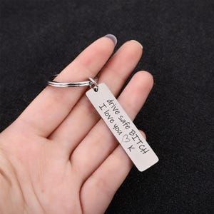 May include: A silver rectangular keyring with the text "drive safe BITCH I love you K" engraved on it. The keyring is attached to a silver ring.