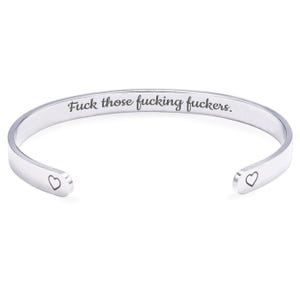 May include: A silver-colored cuff bracelet with the phrase "Fuck those fucking fuckers" engraved on the outside. The open ends of the bracelet each feature a small heart-shaped design. The bracelet is displayed against a white background.