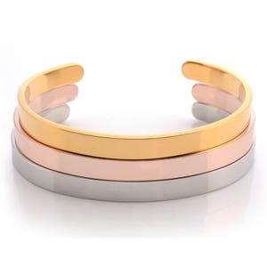 May include: A set of three stacked cuff bracelets featuring a smooth, polished finish. The bracelets are crafted in three distinct metallic tones: a bright yellow gold, a soft rose gold, and a cool, reflective silver, each with an open back.