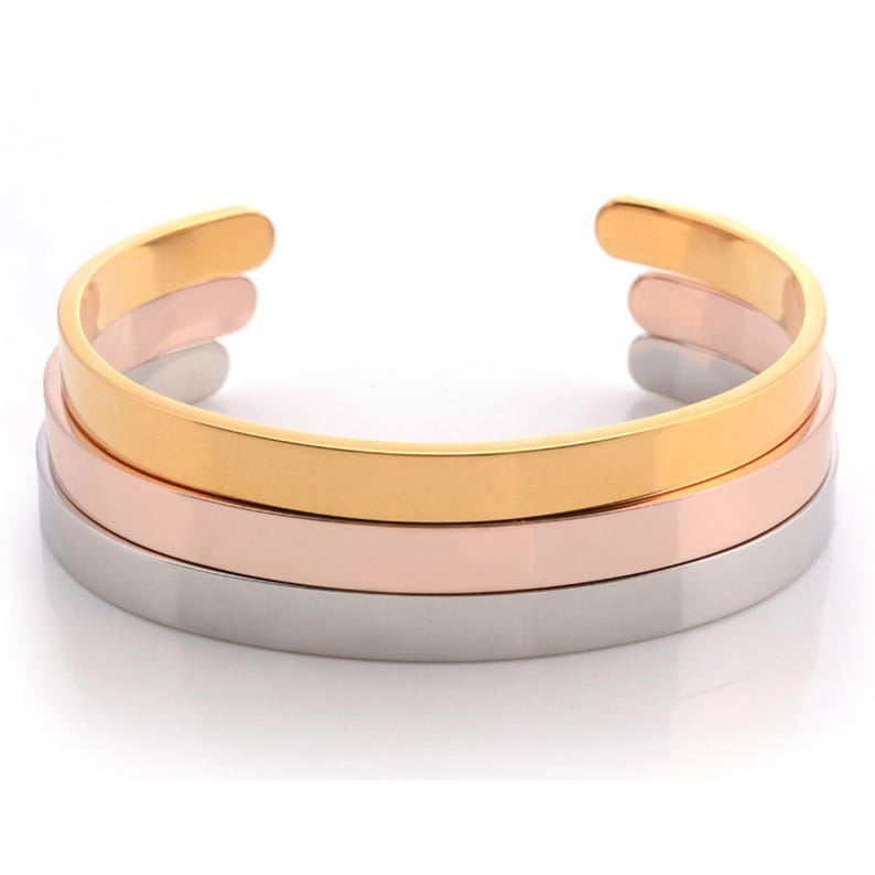 May include: A set of three stacked cuff bracelets featuring a smooth, polished finish. The bracelets are crafted in three distinct metallic tones: a bright yellow gold, a soft rose gold, and a cool, reflective silver, each with an open back.