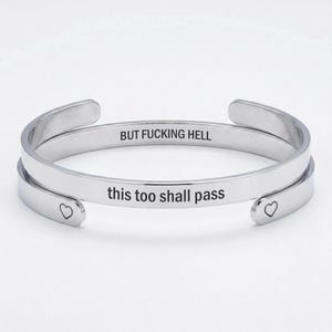 May include: A silver-toned metal cuff bracelet with an open design. The outer surface is engraved with the text "this too shall pass" and features a small heart symbol at each end. The inner surface is engraved with the text "BUT FUCKING HELL".