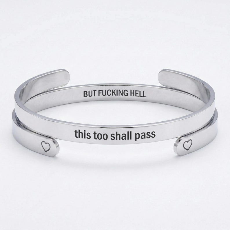 May include: A silver-toned metal cuff bracelet with an open design. The outer surface is engraved with the text "this too shall pass" and features a small heart symbol at each end. The inner surface is engraved with the text "BUT FUCKING HELL".