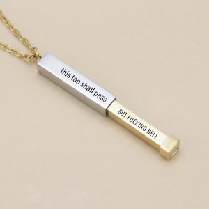 May include: A silver and gold-coloured rectangular pendant necklace. The pendant is engraved with the phrases "this too shall pass" and "BUT FUCKING HELL". The necklace has a gold-coloured chain.