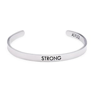 May include: A silver-colored cuff bracelet with the word "STRONG" engraved on the front and "AS FUCK" on the end. The bracelet has a simple, minimalist design.