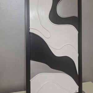 May include: Abstract wall art in a black frame. The artwork features a layered design with black, white, and gray curved shapes. The piece has a modern aesthetic, suitable for home decor. The frame is rectangular.