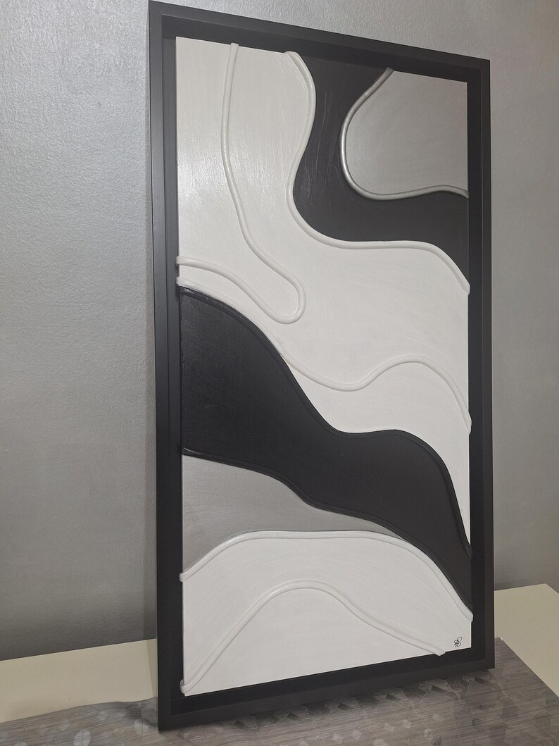 May include: Abstract wall art in a black frame. The artwork features a layered design with black, white, and gray curved shapes. The piece has a modern aesthetic, suitable for home decor. The frame is rectangular.