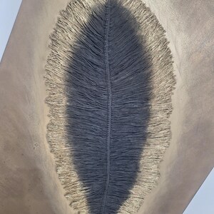 May include: A close-up view of a gray, leaf-shaped macrame wall hanging. The central portion is a darker gray, with a lighter, fringed border. The background is a neutral, light brown color, creating a contrast that highlights the texture and design of the macrame.