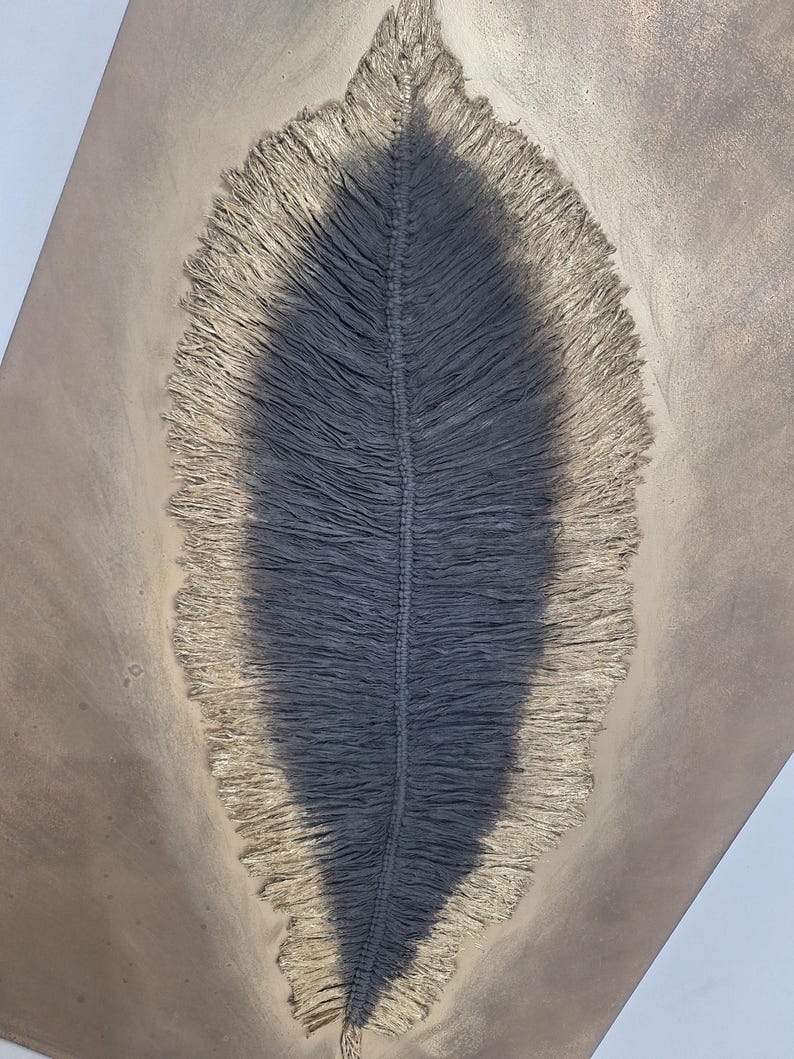 May include: A close-up view of a gray, leaf-shaped macrame wall hanging. The central portion is a darker gray, with a lighter, fringed border. The background is a neutral, light brown color, creating a contrast that highlights the texture and design of the macrame.