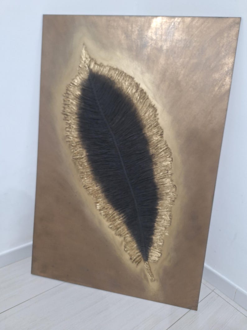 May include: A rectangular wall art piece featuring a large, stylized feather design. The feather's center is black, surrounded by a textured gold border, set against a bronze-colored background. The artwork is mounted on a wooden frame.