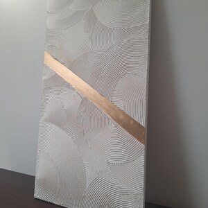 May include: Textured white rectangular wall art with a gold diagonal stripe. The white surface features a repeating pattern of curved lines, creating a layered, circular design. The gold stripe adds a contrasting metallic accent to the artwork.