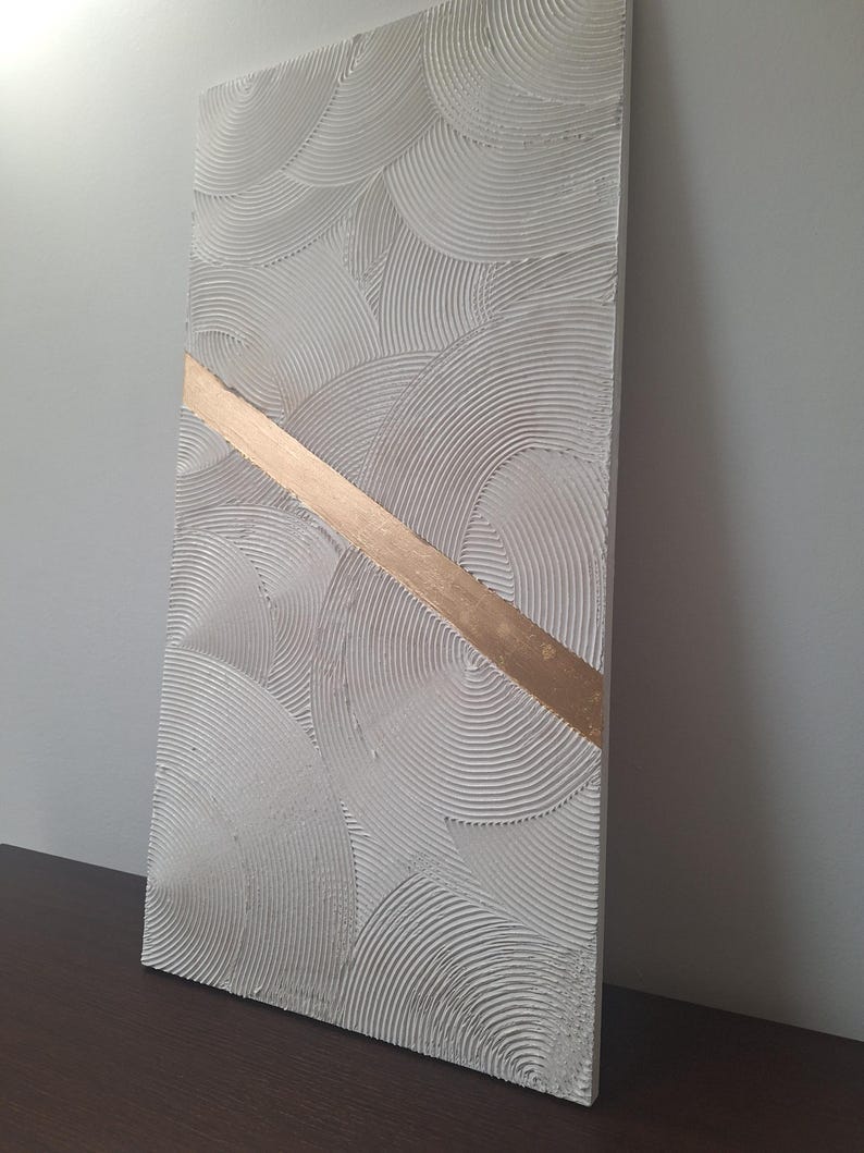 May include: Textured white rectangular wall art with a gold diagonal stripe. The white surface features a repeating pattern of curved lines, creating a layered, circular design. The gold stripe adds a contrasting metallic accent to the artwork.