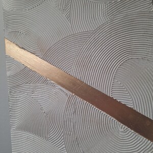 May include: Textured abstract art piece with a gray background and a diagonal copper-colored stripe. The gray surface features a repeating pattern of curved lines, creating a sense of depth and movement. This artwork is suitable for home decor.