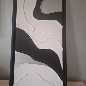 May include: Abstract wall art in a rectangular black frame. The artwork features a layered design with flowing shapes in shades of white, gray, and black. The piece has a modern, minimalist aesthetic, suitable for home decor.