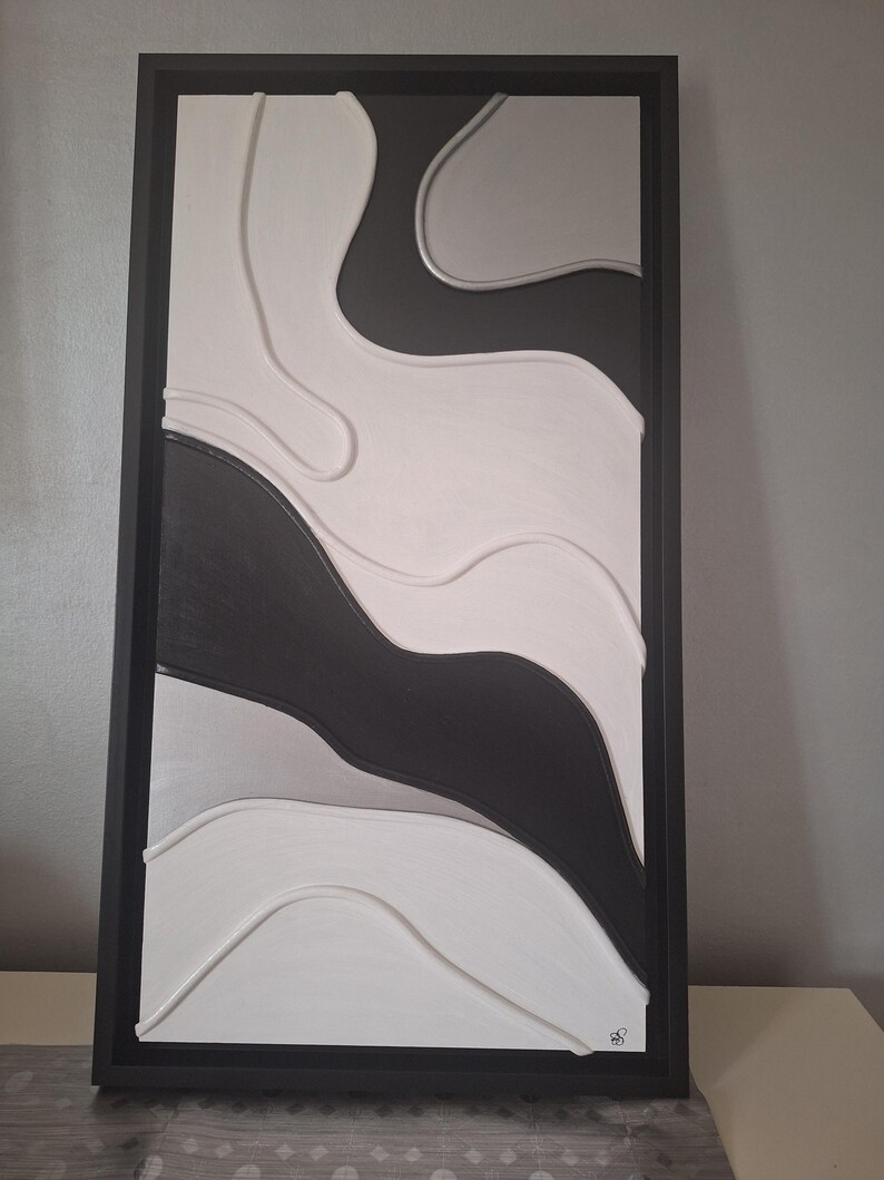 May include: Abstract wall art in a rectangular black frame. The artwork features a layered design with flowing shapes in shades of white, gray, and black. The piece has a modern, minimalist aesthetic, suitable for home decor.