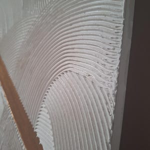 May include: Close-up of a textured, white surface with curved, parallel lines. A vertical, light brown strip bisects the design. The texture appears to be applied with a trowel, creating a wave-like pattern.