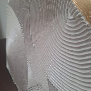 May include: Close-up of a textured, off-white artwork with a wave-like pattern. The edge of the piece features a gold leaf finish, adding a touch of elegance. The surface has a tactile, layered appearance.
