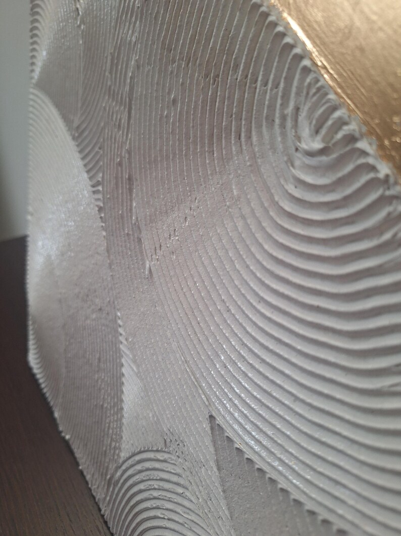 May include: Close-up of a textured, off-white artwork with a wave-like pattern. The edge of the piece features a gold leaf finish, adding a touch of elegance. The surface has a tactile, layered appearance.