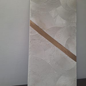 May include: Textured white wall art with a repeating circular pattern and a diagonal gold-colored stripe. The artwork is rectangular and appears to be made of plaster or a similar material. The piece is displayed against a neutral-colored wall.