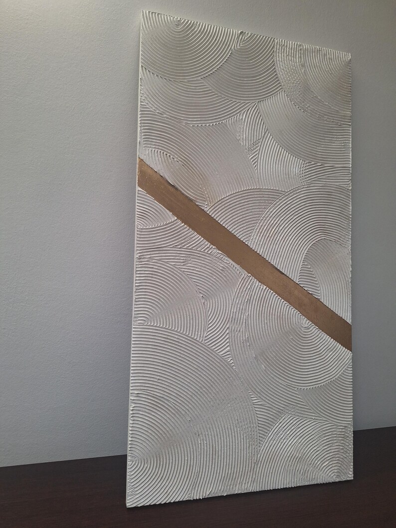 May include: Textured white wall art with a repeating circular pattern and a diagonal gold-colored stripe. The artwork is rectangular and appears to be made of plaster or a similar material. The piece is displayed against a neutral-colored wall.