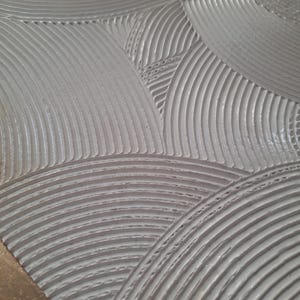 May include: Close-up of a textured, light gray surface with a repeating pattern of curved lines and circular shapes. The design has a three-dimensional effect, with the lines creating a sense of depth and texture. The overall aesthetic is modern and minimalist.