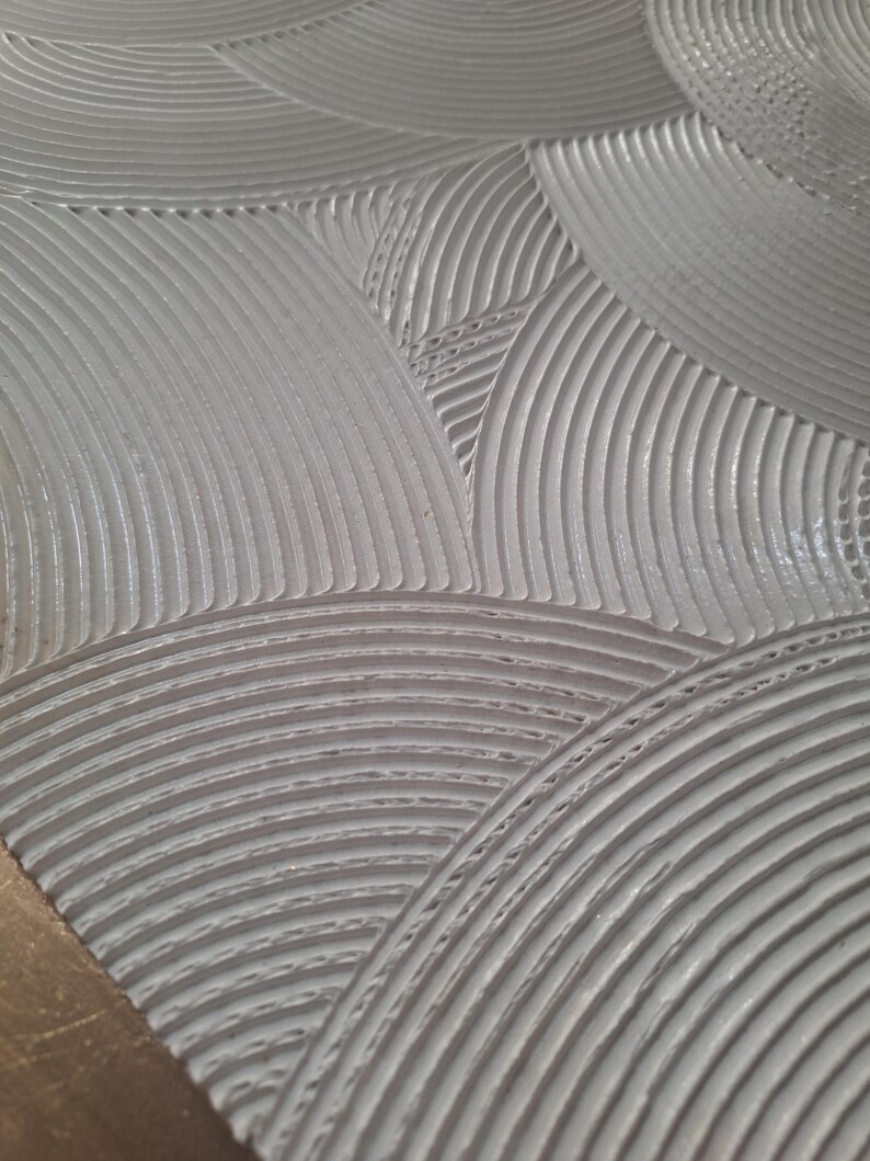 May include: Close-up of a textured, light gray surface with a repeating pattern of curved lines and circular shapes. The design has a three-dimensional effect, with the lines creating a sense of depth and texture. The overall aesthetic is modern and minimalist.