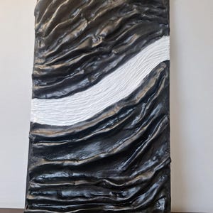 May include: Abstract wall art featuring a textured, rectangular panel. The artwork is predominantly black with a flowing, rippled design. A white, textured stripe bisects the panel diagonally, creating a striking contrast. The piece has a modern, sculptural feel.