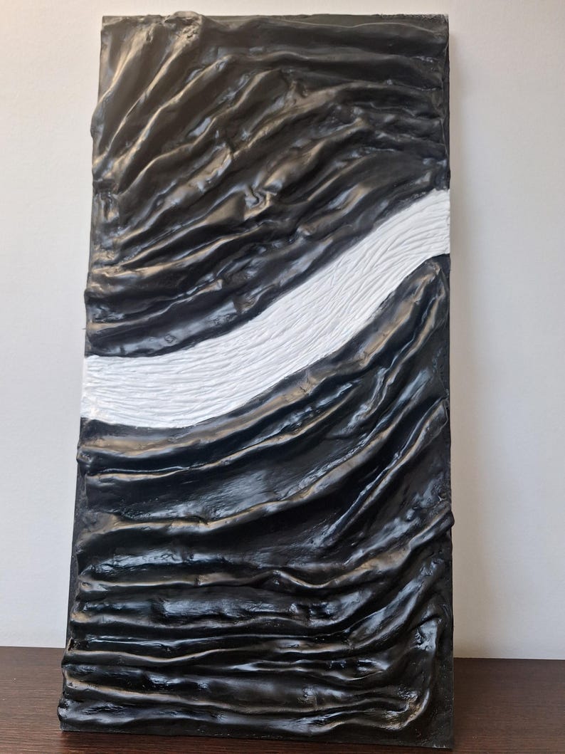 May include: Abstract wall art featuring a textured, rectangular panel. The artwork is predominantly black with a flowing, rippled design. A white, textured stripe bisects the panel diagonally, creating a striking contrast. The piece has a modern, sculptural feel.
