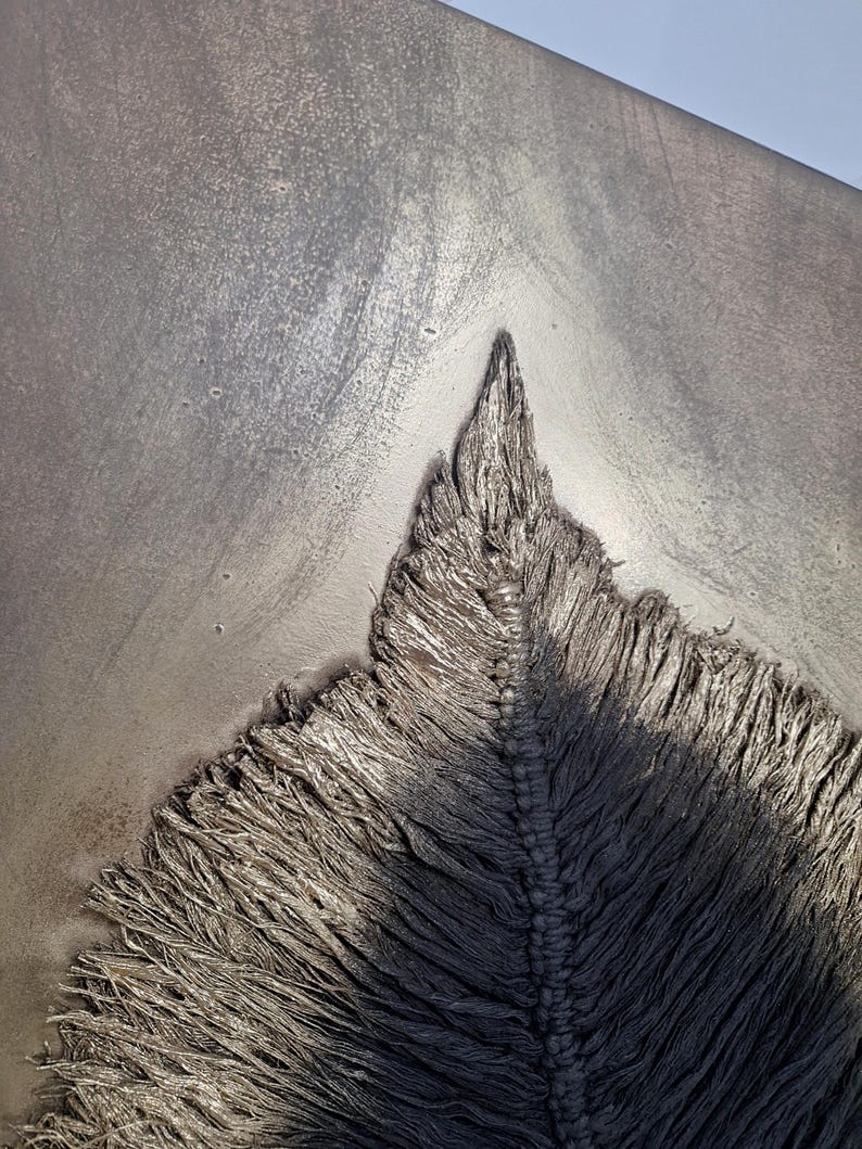 May include: Close-up of a textured, gray artwork with a central, pointed design. The artwork features a rough, frayed texture, with a central ridge and a gradient of light and shadow.