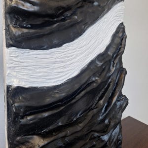 May include: A textured, abstract sculpture with a striking contrast of black and white. The artwork features a flowing, wave-like white section that cuts through the predominantly black, rough-hewn surface. The piece has a modern, artistic aesthetic.