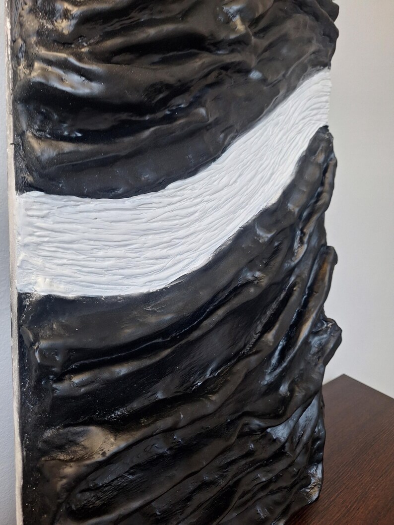 May include: A textured, abstract sculpture with a striking contrast of black and white. The artwork features a flowing, wave-like white section that cuts through the predominantly black, rough-hewn surface. The piece has a modern, artistic aesthetic.