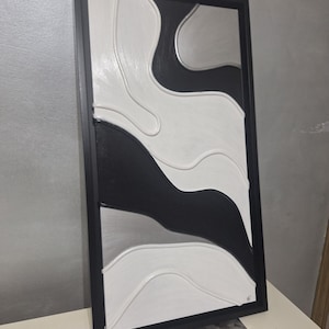May include: Abstract wall art in a black frame. The artwork features a textured design with flowing shapes in black, white, and silver. The piece is a modern, minimalist design suitable for home decor.