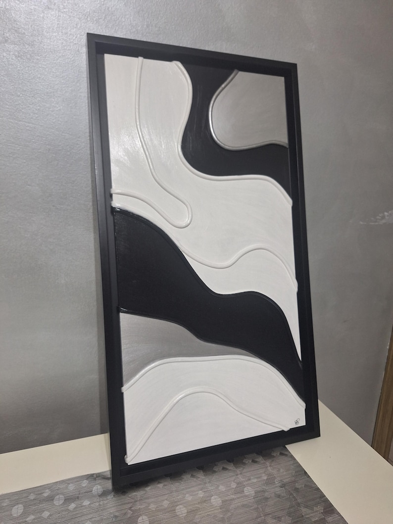 May include: Abstract wall art in a black frame. The artwork features a textured design with flowing shapes in black, white, and silver. The piece is a modern, minimalist design suitable for home decor.