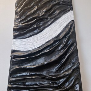 May include: Abstract wall art featuring a textured, rectangular panel. The artwork is predominantly black with a central white stripe. The surface has a rippled, wave-like appearance, creating a sense of depth and movement. The piece is mounted on a white wall.