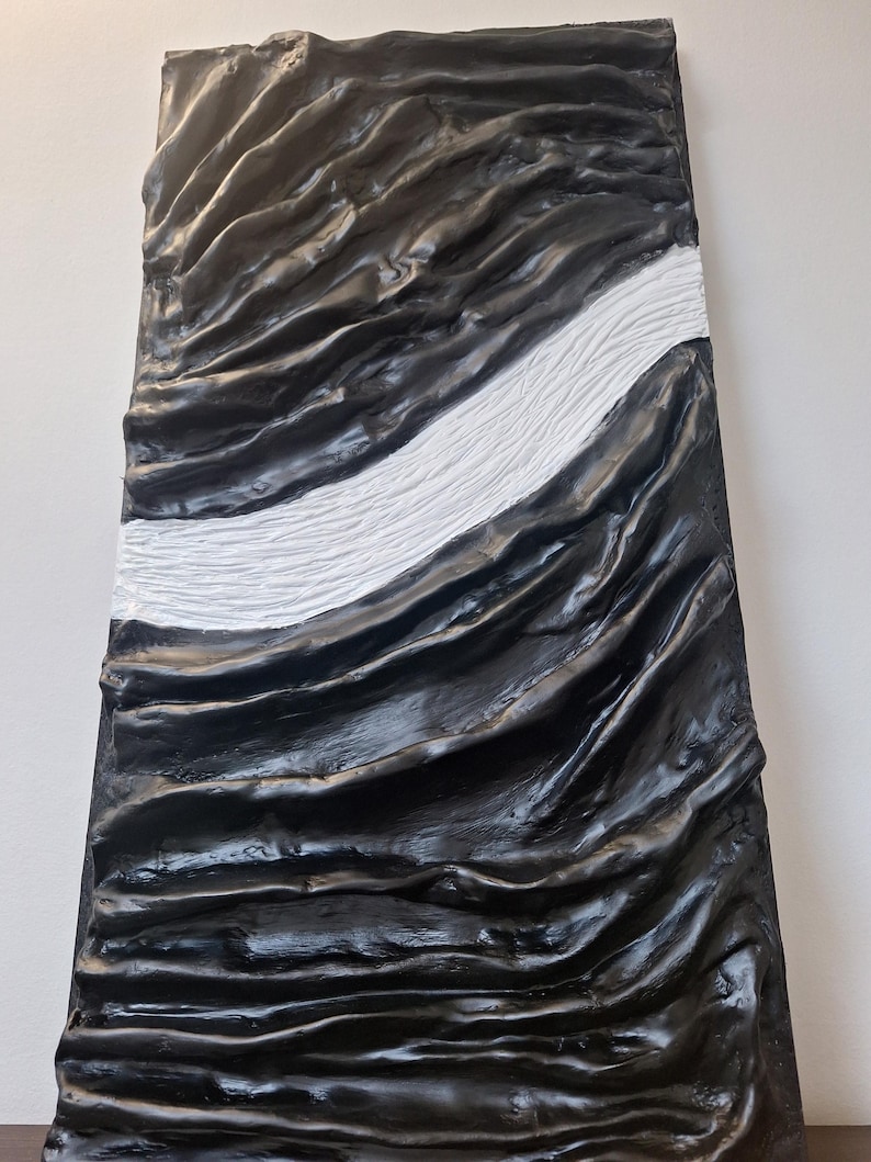 May include: Abstract wall art featuring a textured, rectangular panel. The artwork is predominantly black with a central white stripe. The surface has a rippled, wave-like appearance, creating a sense of depth and movement. The piece is mounted on a white wall.