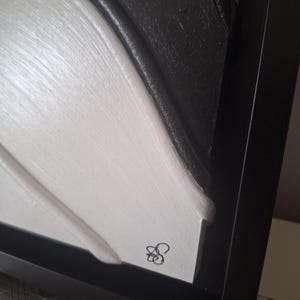 May include: Abstract framed artwork with a textured surface. The piece features a contrast of black and white, with a signature in the lower right corner. The artwork is encased in a black frame.