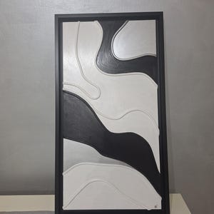 May include: Abstract wall art in a black frame. The artwork features a layered design with black, white, and gray curved shapes. The piece is mounted on a white surface against a gray wall.