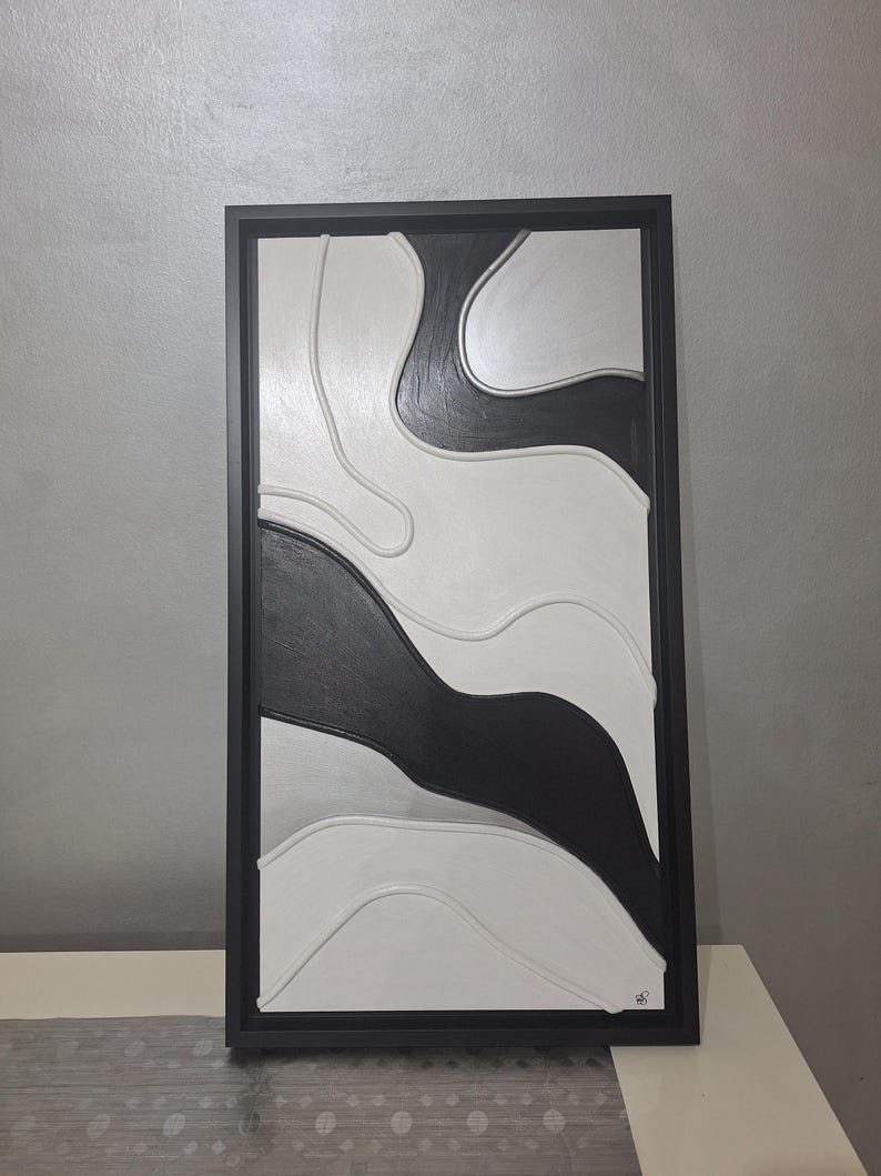 May include: Abstract wall art in a black frame. The artwork features a layered design with black, white, and gray curved shapes. The piece is mounted on a white surface against a gray wall.