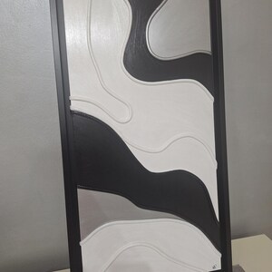 May include: Abstract wall art in a black frame. The artwork features a layered design with flowing shapes in black, white, and gray. The piece has a textured appearance, adding depth and visual interest to the modern design.