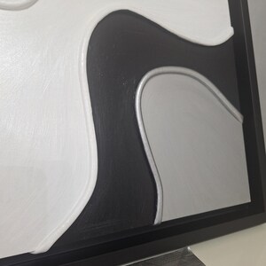 May include: Abstract wall art in a black frame. The artwork features a textured design with white, black, and silver-gray curved shapes. The piece has a modern, minimalist aesthetic, suitable for various interior styles.