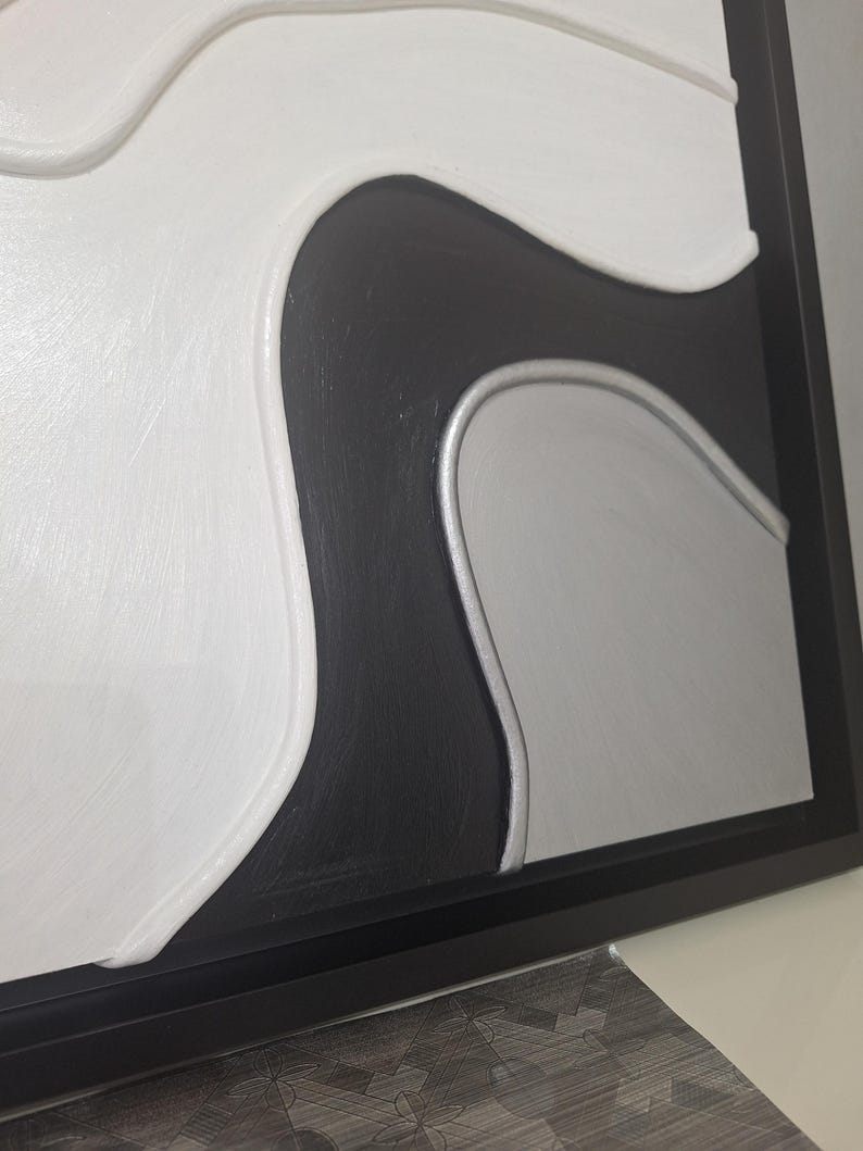 May include: Abstract wall art in a black frame. The artwork features a textured design with white, black, and silver-gray curved shapes. The piece has a modern, minimalist aesthetic, suitable for various interior styles.