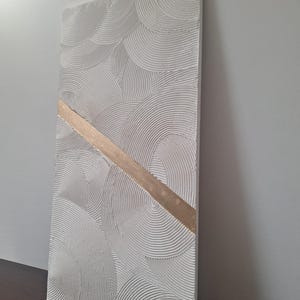 May include: Textured white rectangular wall art with a repeating semi-circular pattern. A diagonal gold-colored stripe bisects the artwork. The piece is displayed against a neutral background, showcasing its three-dimensional texture.
