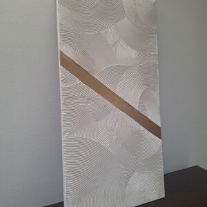 May include: Textured white rectangular wall art with a diagonal gold stripe. The white surface features a repeating pattern of curved lines, creating a layered, circular design. The artwork is displayed on a dark brown surface.