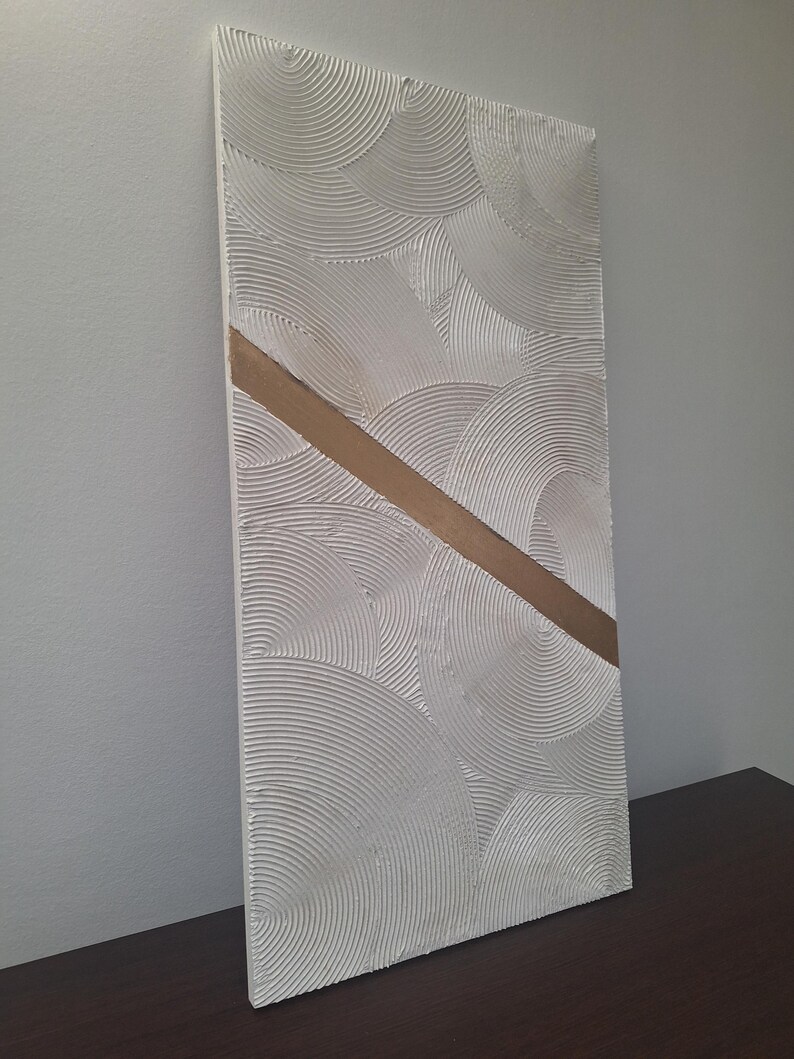 May include: Textured white rectangular wall art with a diagonal gold stripe. The white surface features a repeating pattern of curved lines, creating a layered, circular design. The artwork is displayed on a dark brown surface.
