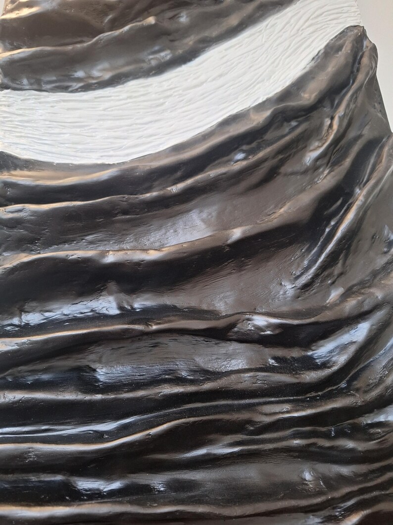 May include: Abstract sculpture with a textured surface. The artwork features flowing, wave-like forms in glossy black, contrasted by a white, textured section. The piece has a dynamic, organic feel.